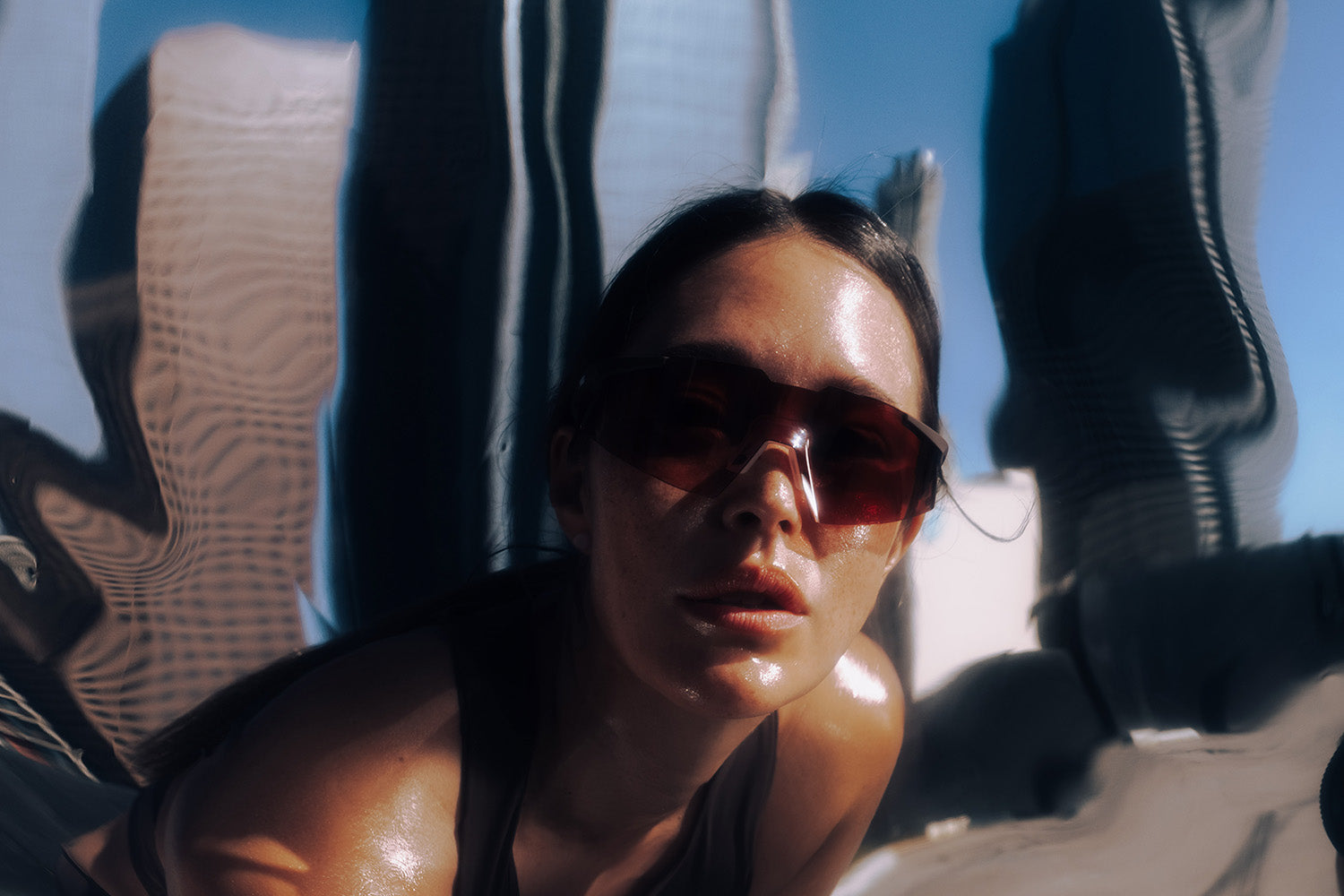 Female runner reclining and looking upward in harsh sunlight, wearing 100Percent® A2 sunglasses, glass city towers reflected in the frame behind her.