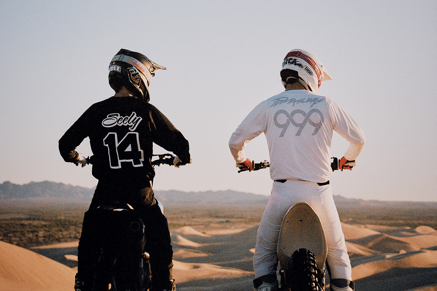 Two riders in 100Percent® goggles sit on their bikes atop a dune at dusk, one in a black number 14 jersey and one in white, overlooking an endless sea of Glamis dunes.