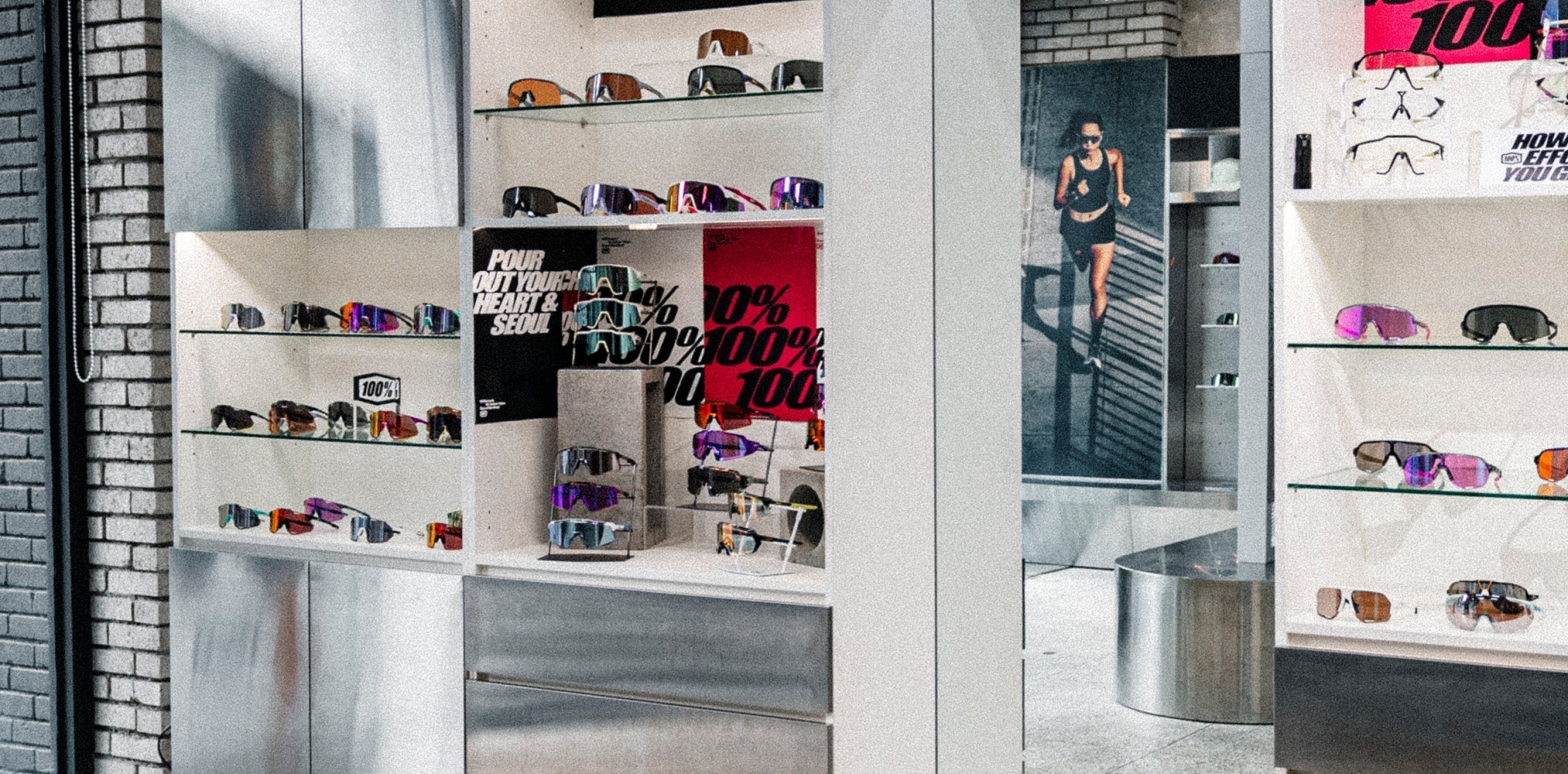 100Percent® Seoul, South Korea Marathon Pop Up. A retail display of various 100% sunglasses on white shelving units inside a modern store, with a brick wall on the left and a large 100% promotional poster in the background.