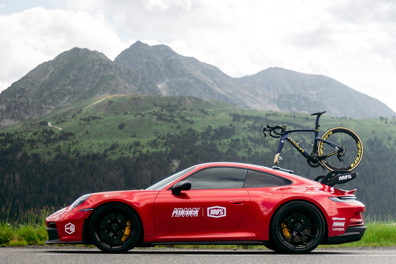 Red Porsche 911 GT3 RS wrapped in Tom Pidcock x 100Percent® livery with a Pinarello road bike mounted on the roof rack, parked against a mountain backdrop near the Nürburgring