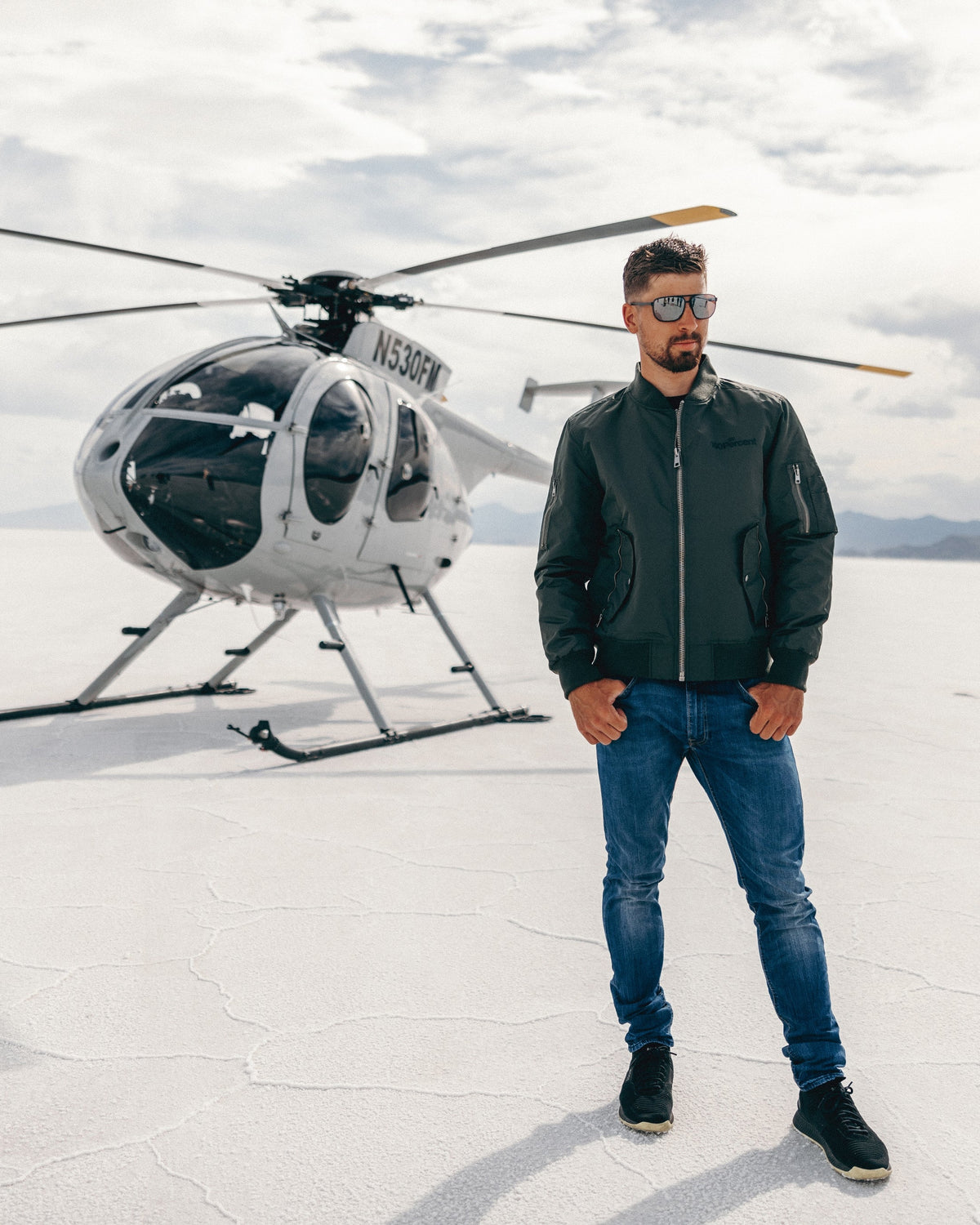 Peter Sagan wearing a black bomber jacket, blue jeans, and black sneakers stands in front of a white helicopter on a flat, cracked white desert surface under a cloudy sky.