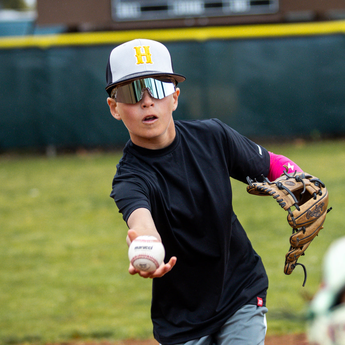 A Person in a baseball cap and 100Percent sunglasses throwing a baseball and glove on a field
